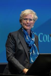 A blonde woman wearing a blue scarf stands at a podium